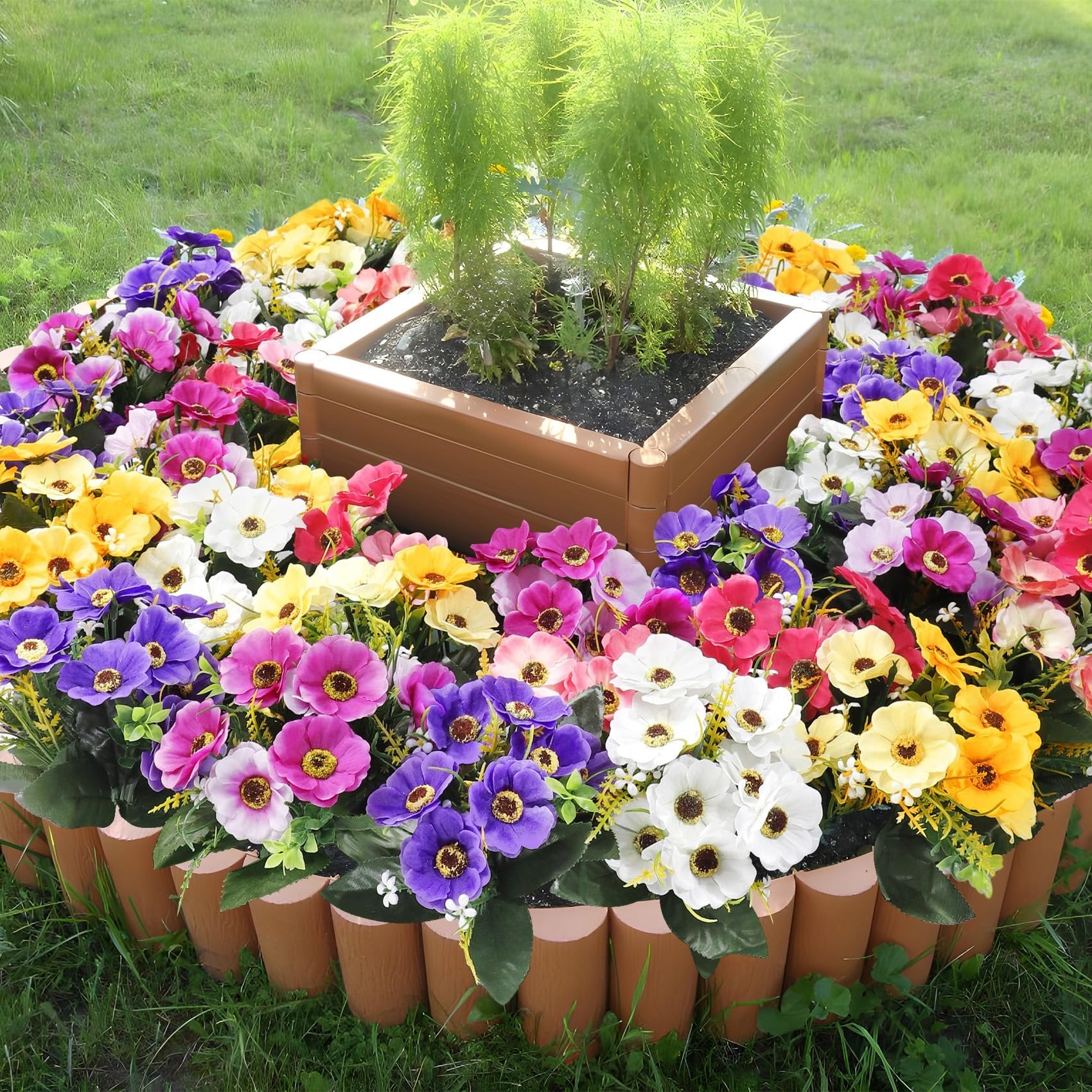 Circular arrangement of mixed color artificial daisy silk flowers around a planter with green foliage.