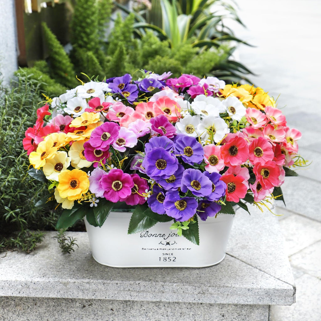 Mixed color artificial daisy flowers in a white planter with text, set outdoors on a stone surface.