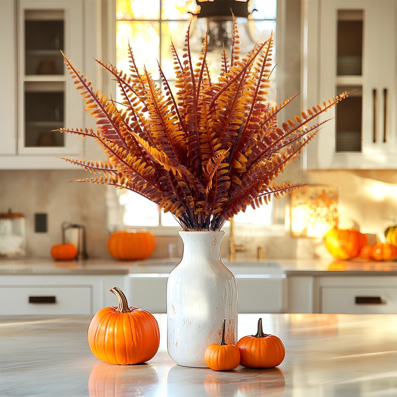 Autumnal faux fern arrangement in a white vase with pumpkins on a kitchen counter, with a window in the background.