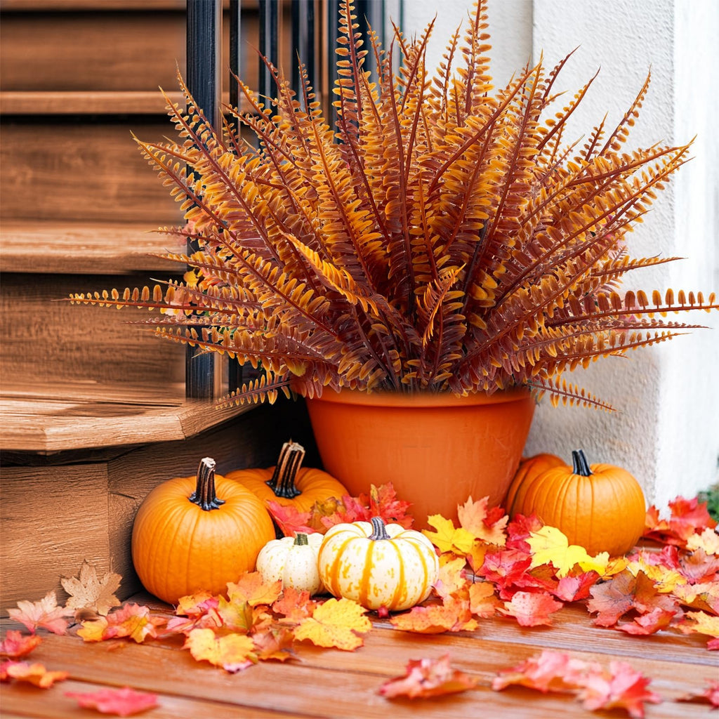 Autumnal display of artificial fern in an orange pot, surrounded by pumpkins and colorful fall leaves on a wooden porch.