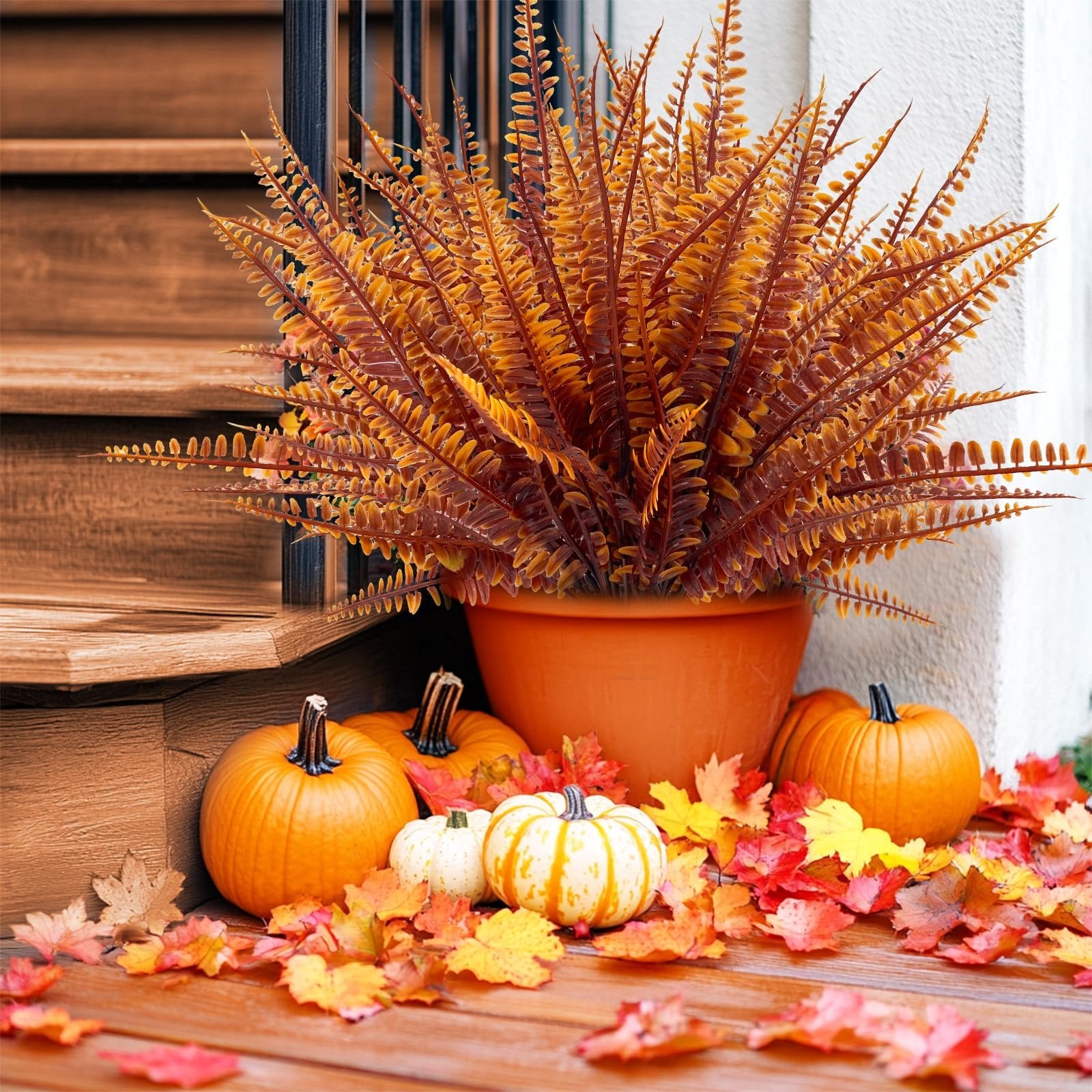 Autumnal display of artificial fern in an orange pot, surrounded by pumpkins and colorful fall leaves on a wooden porch.