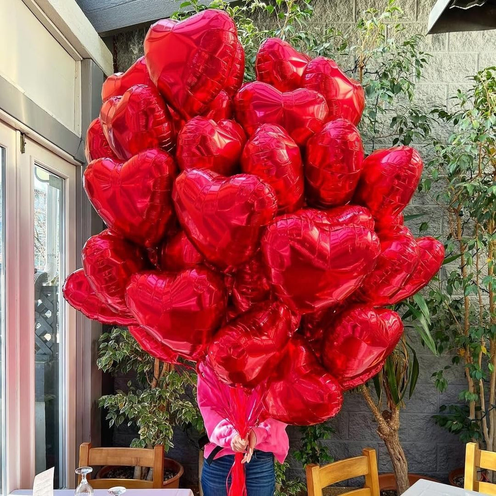 A person holding a large bunch of red heart-shaped balloons outdoors with greenery and tables in the background.
