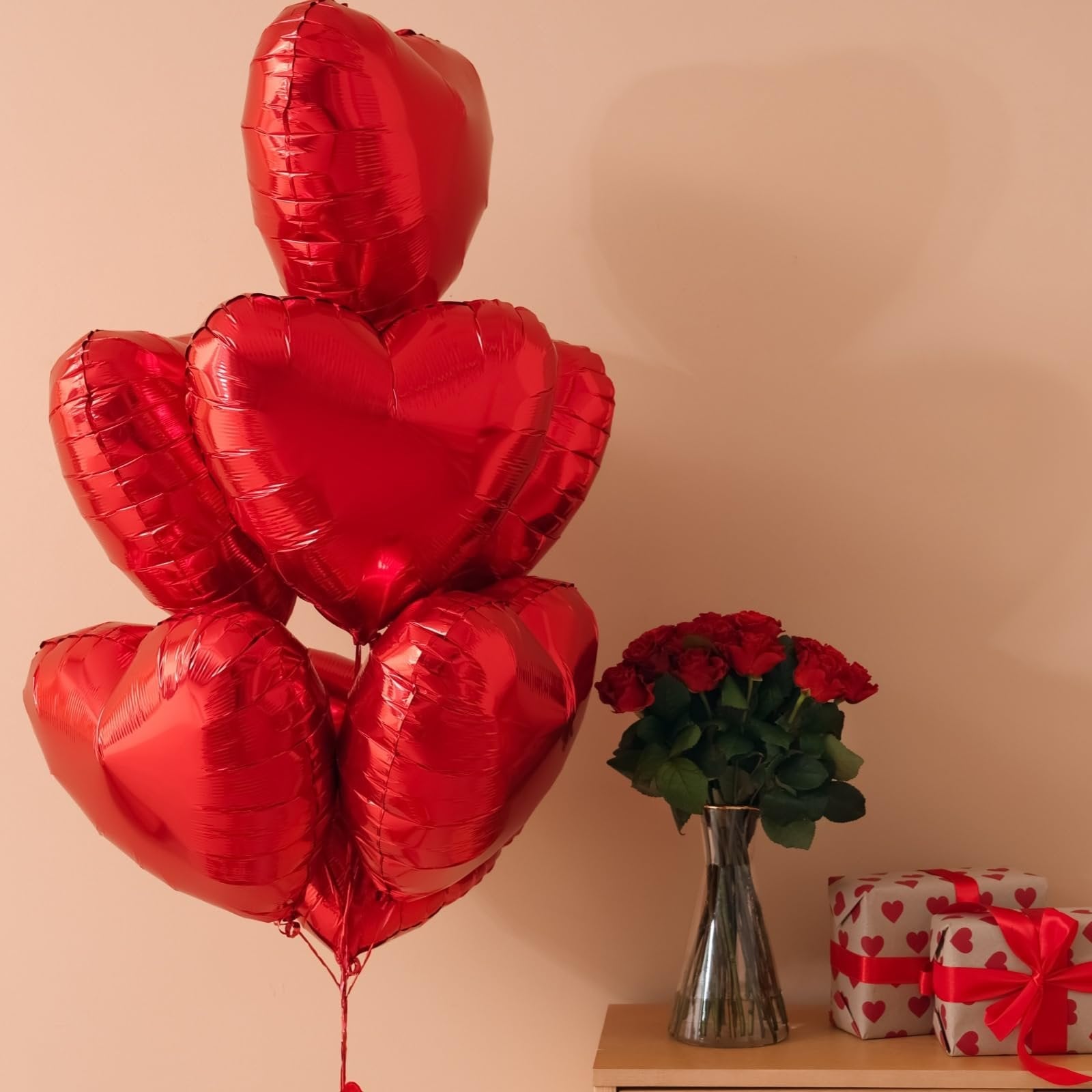 Cluster of red heart-shaped foil balloons with a bouquet of red roses and two gift-wrapped boxes on a shelf.