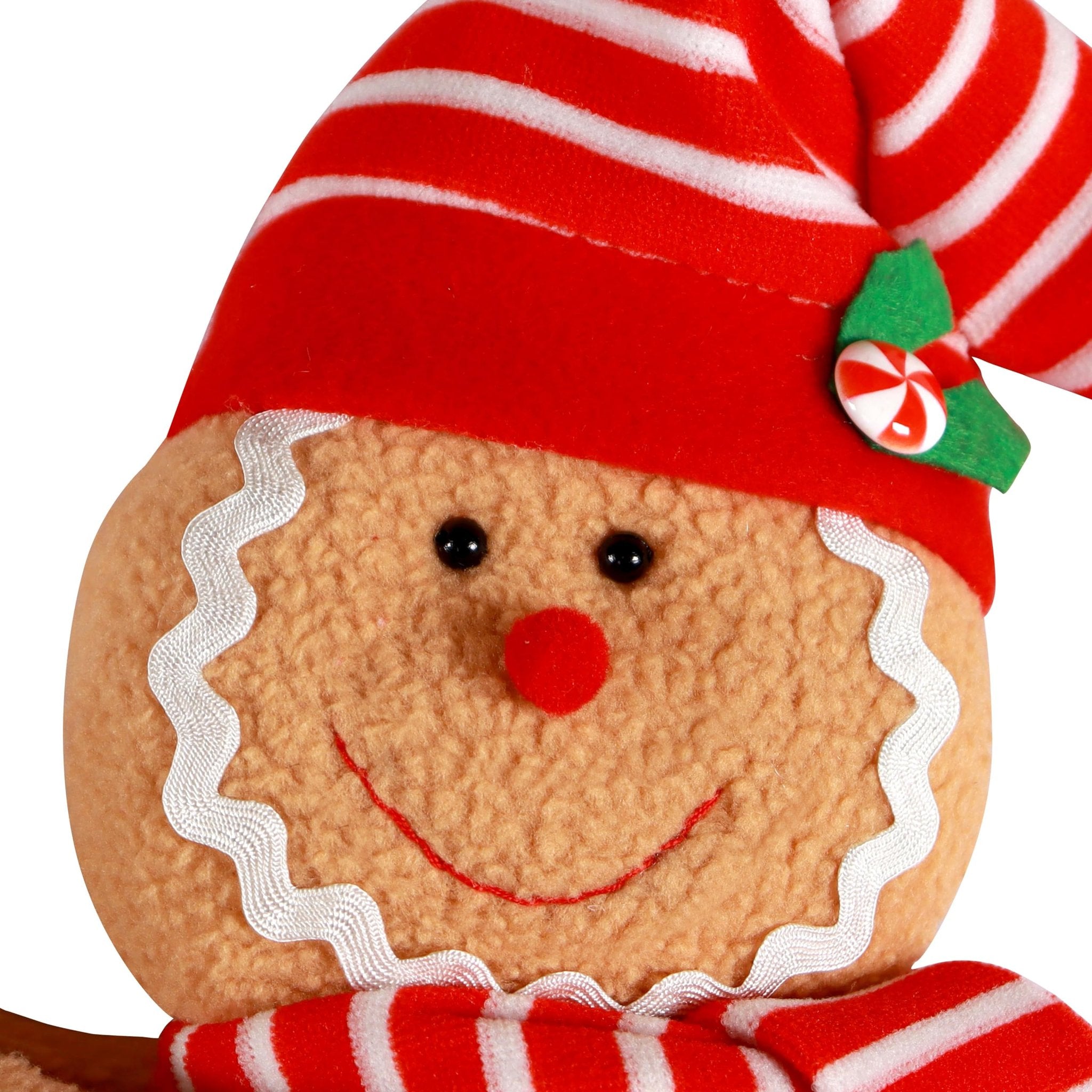 Close-up of a plush gingerbread man Christmas tree topper with a red and white striped hat and scarf, and a candy accent.
