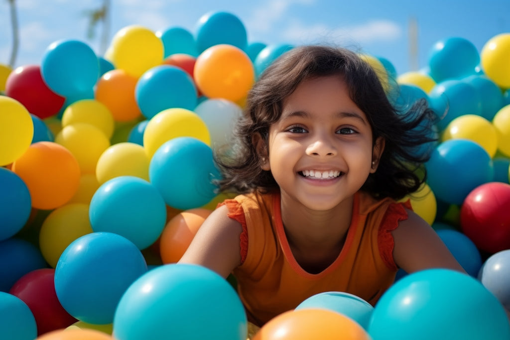 Smiling young girl peeking out from a pile of colorful balloons, including blue, yellow, and orange.