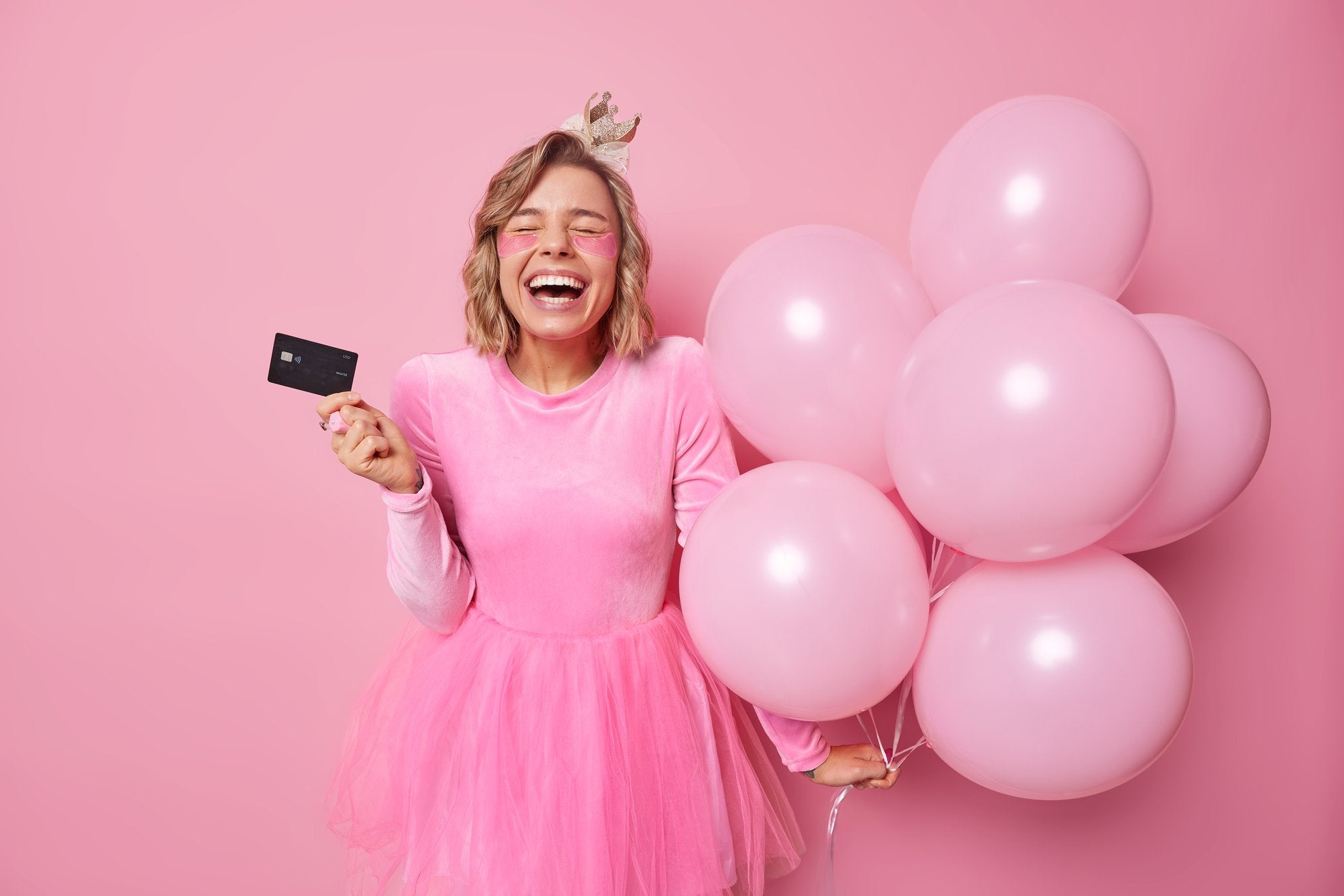 Woman in pink dress and crown laughing, holding credit card and pink balloons