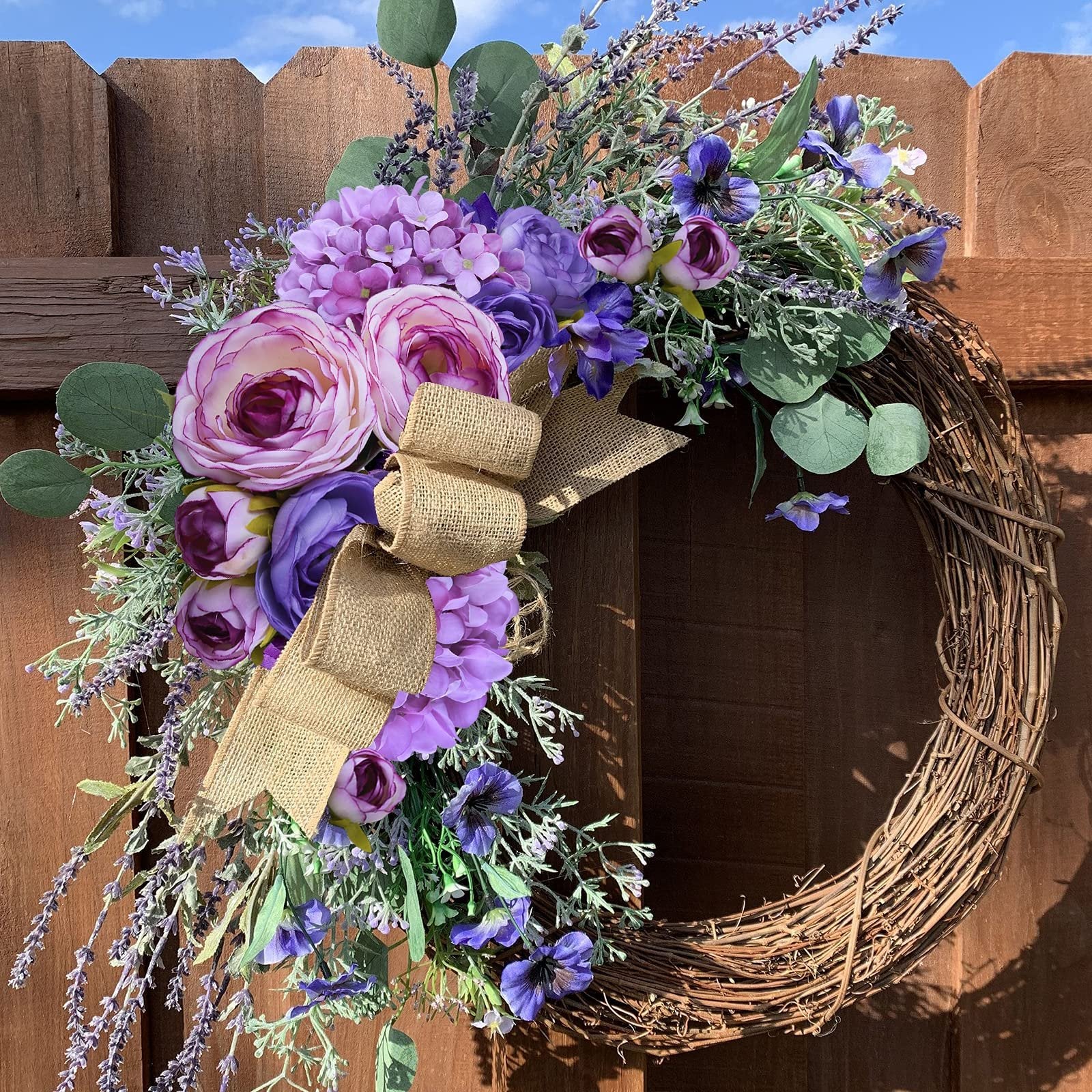 Grapevine wreath adorned with purple silk hydrangea, roses, lavender, and eucalyptus, featuring a burlap bow, hung on a wooden fence.