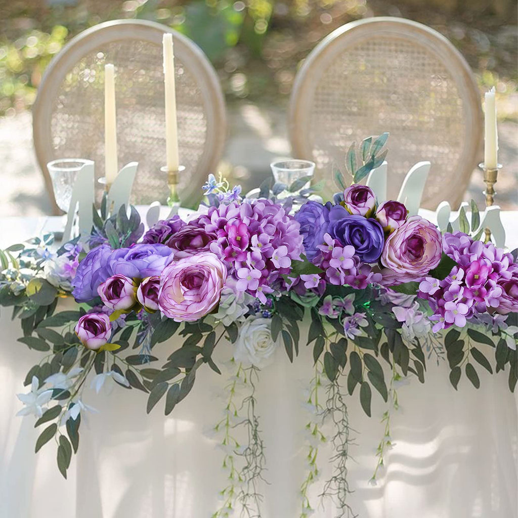Purple and pink silk hydrangea and rose floral arrangement on a draped table with two chairs and candles in the background.