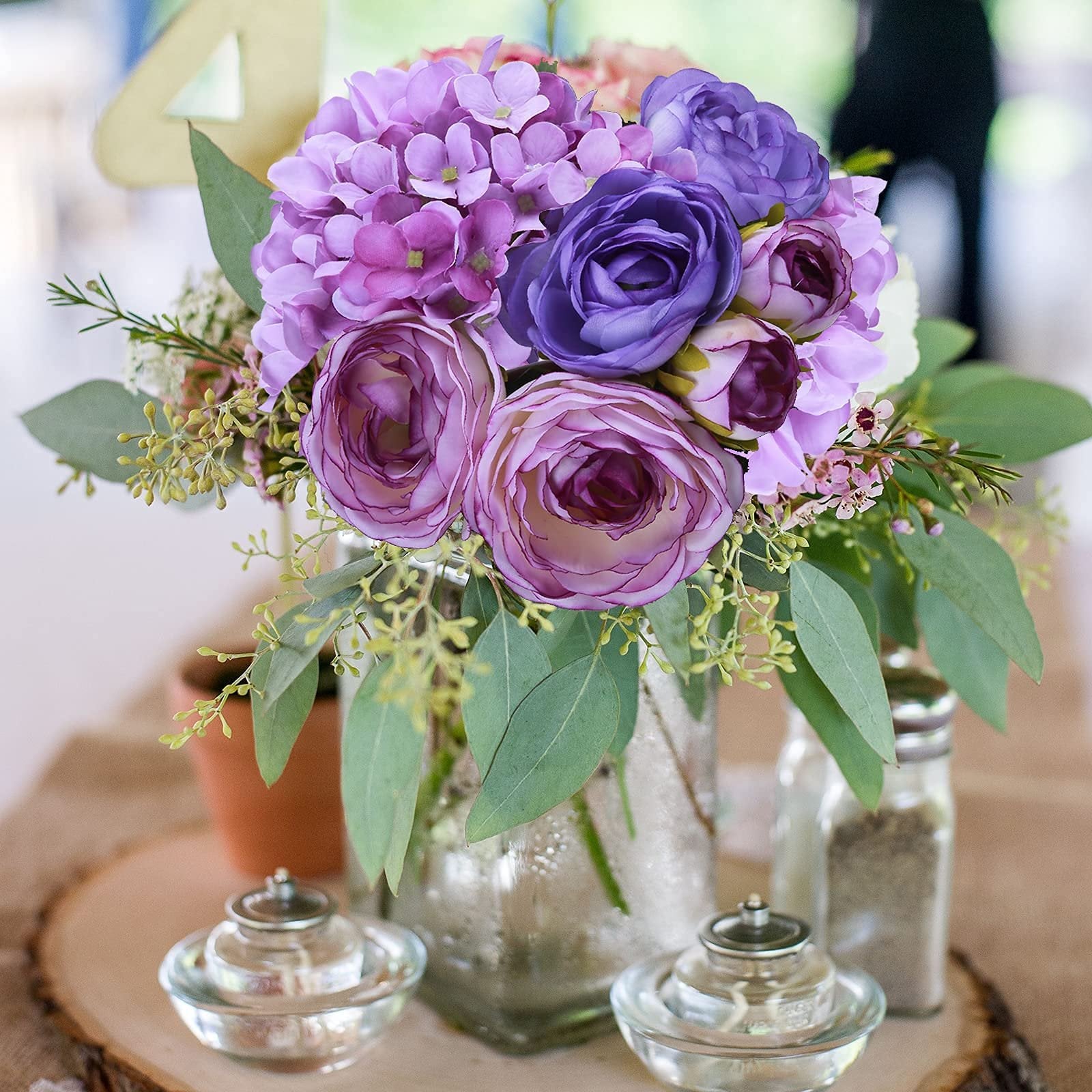 Purple and pink silk hydrangea and rose bouquet in a glass vase on a wooden slice with small oil lamps.