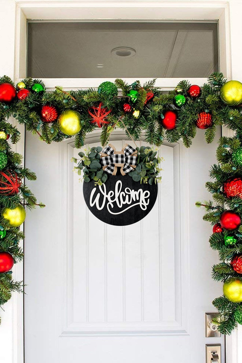 Round black welcome sign with eucalyptus wreath and plaid bow hanging on a white door with Christmas garland