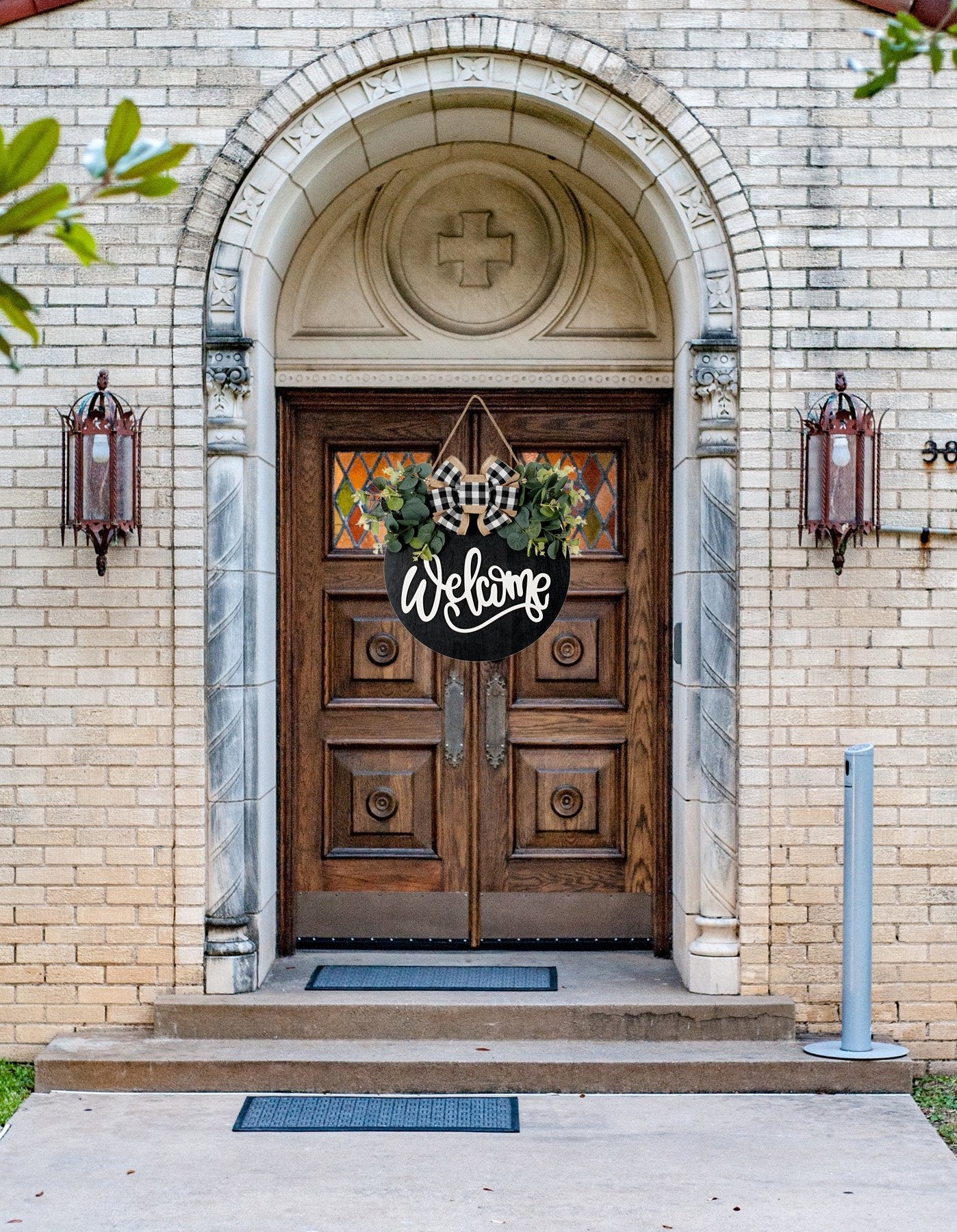 Round wooden welcome sign with eucalyptus wreath and plaid bow hanging on a wooden door.