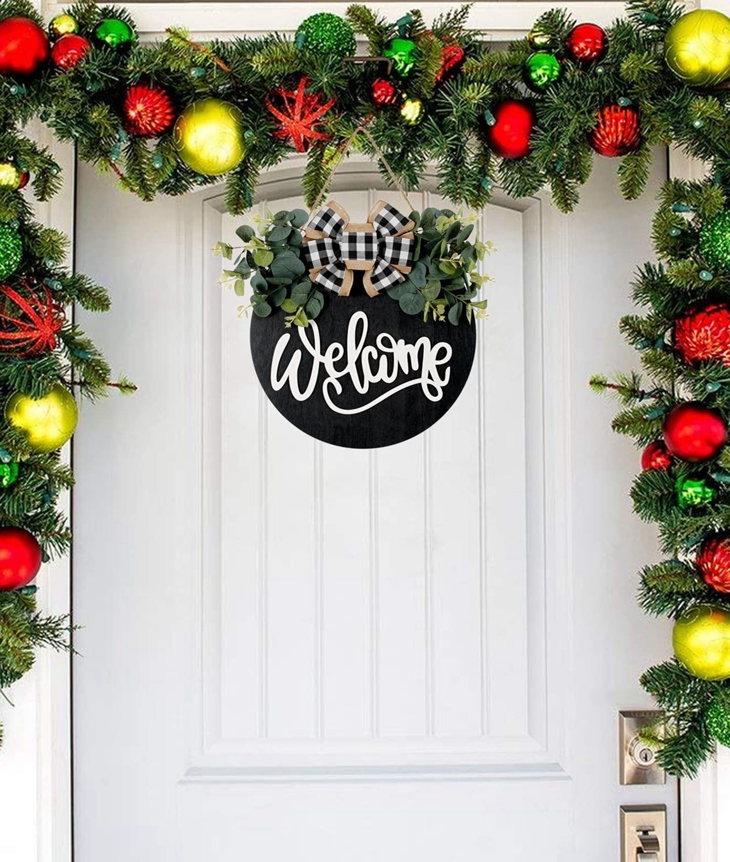Round black welcome sign with eucalyptus and plaid bow hanging on a white door with Christmas garland