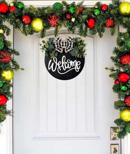 Round black welcome sign with eucalyptus and plaid bow hanging on a white door with Christmas garland