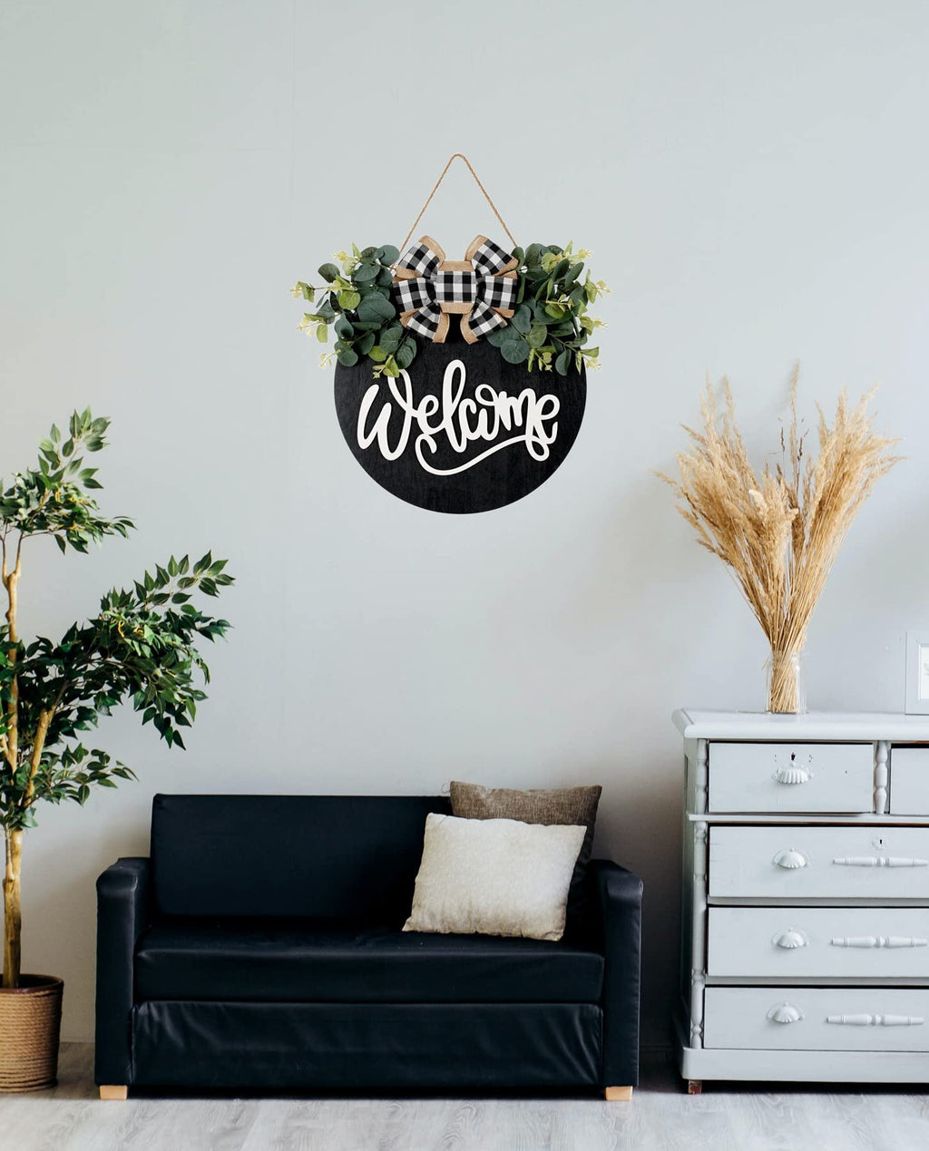 Round black wooden welcome sign with eucalyptus wreath and plaid bow hanging on a wall above a black sofa.