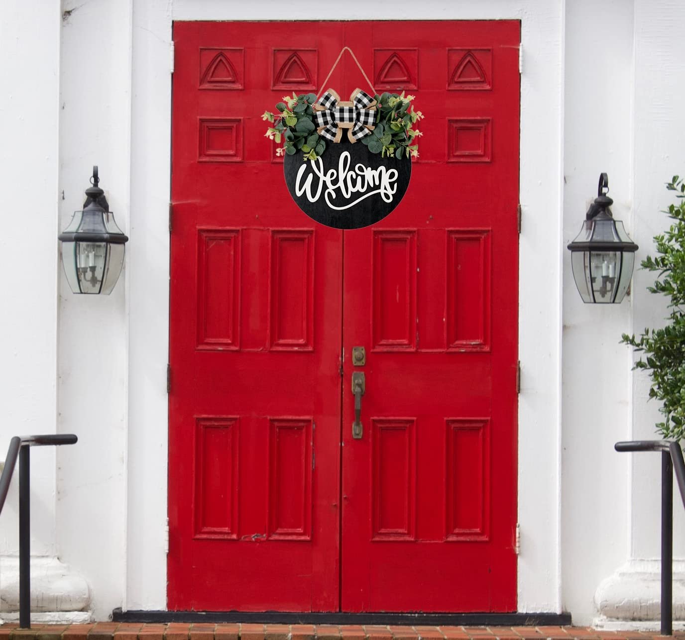 Round wooden welcome sign with eucalyptus wreath and plaid bow hanging on a red door.