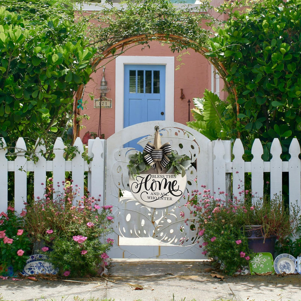 Round wooden welcome sign with floral accents hanging on a white picket fence in front of a pink house.