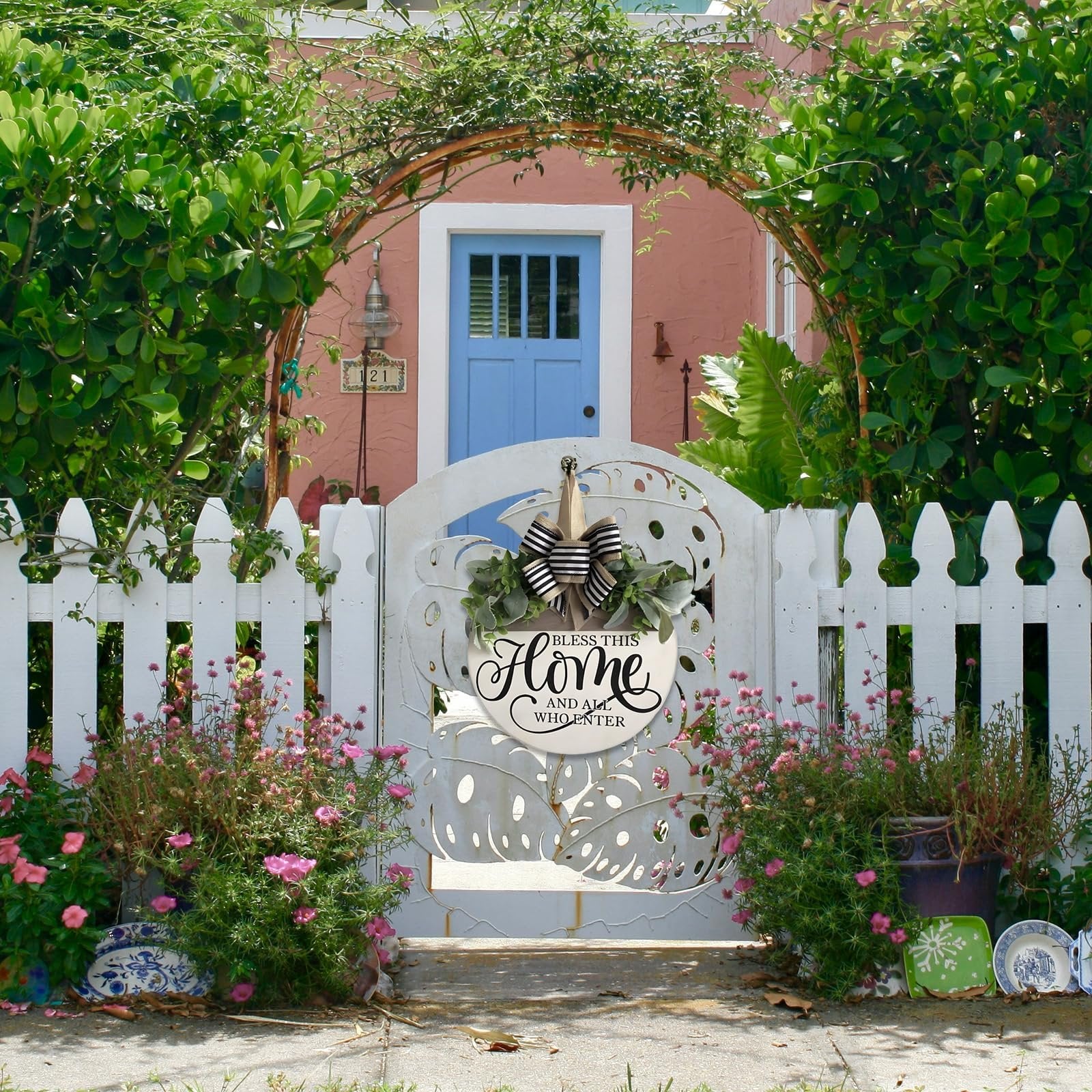 Round wooden welcome sign with floral accents hanging on a white picket fence in front of a pink house.