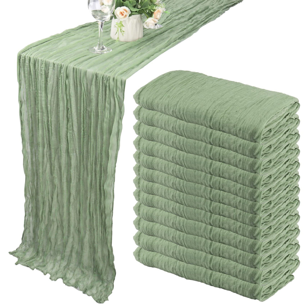 Dusty sage green cheesecloth table runner draped on a table with a wine glass and floral arrangement, next to a stack of 12 folded runners.