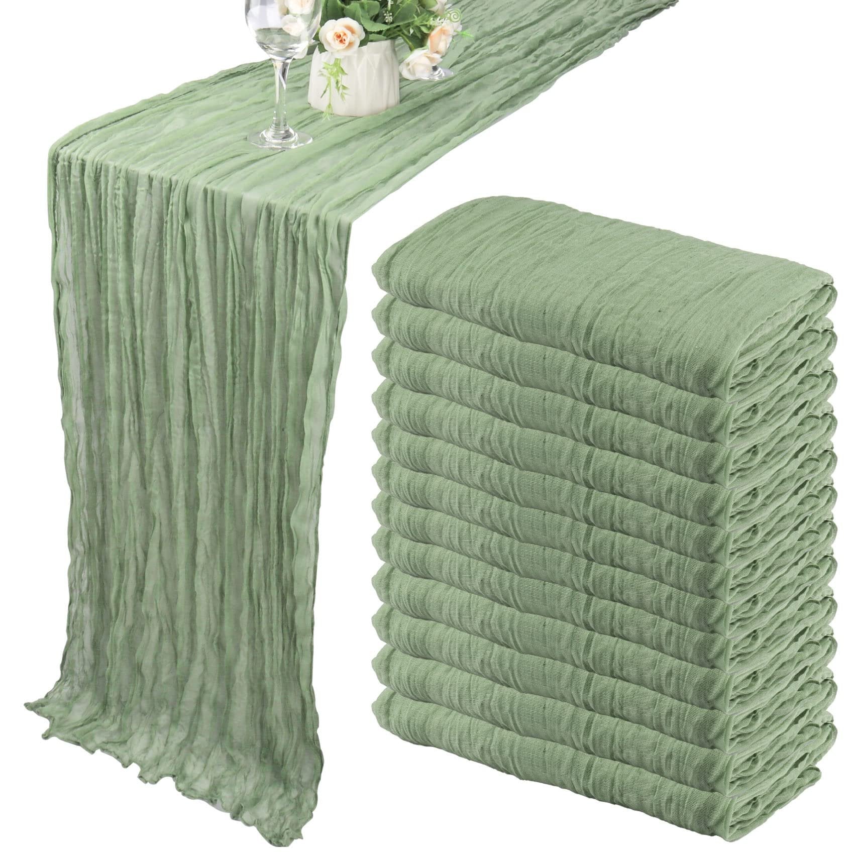 Dusty sage green cheesecloth table runner draped on a table with a wine glass and floral arrangement, next to a stack of 12 folded runners.