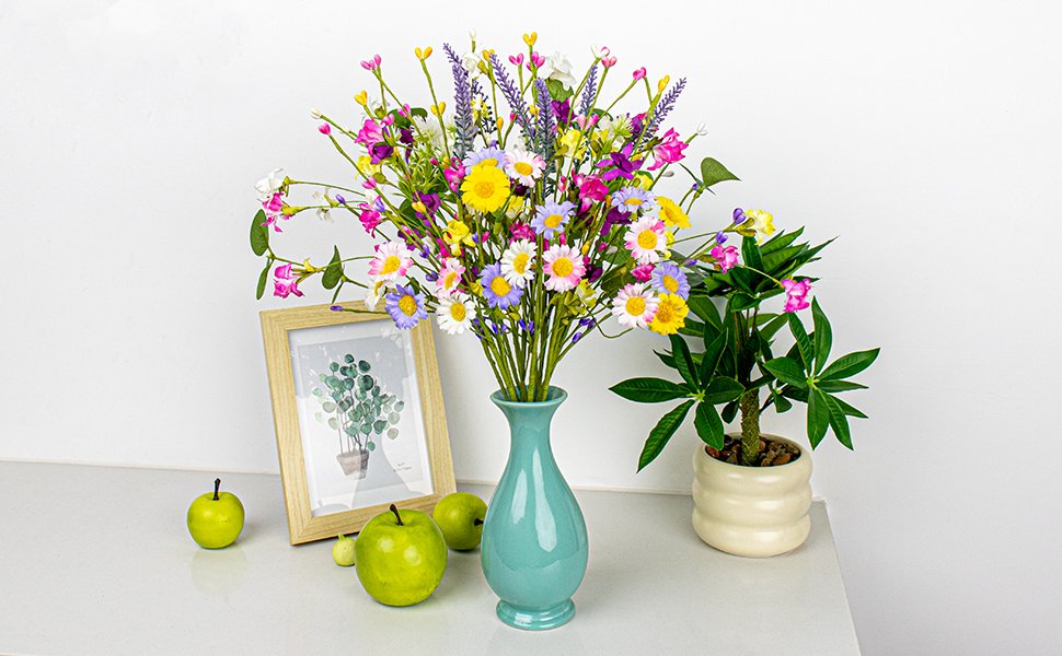 Artificial spring floral arrangement with daisies, wildflowers, and berries in a teal vase, next to a framed picture and green apples.