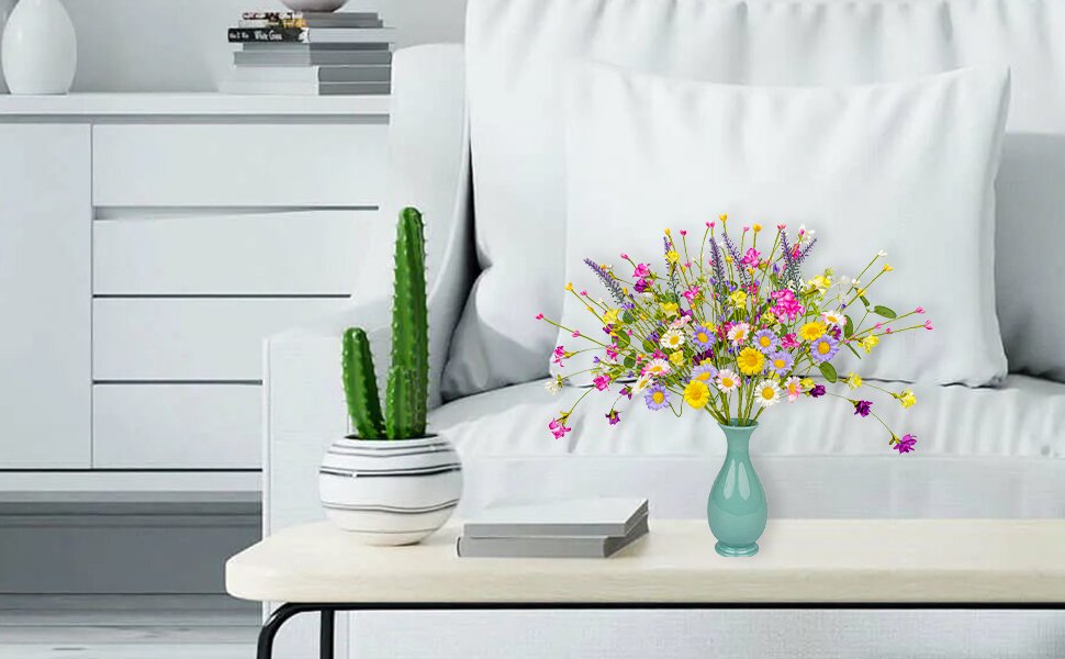 Artificial spring floral arrangement with daisies, wildflowers, and pip berries in a teal vase on a table next to a potted cactus.
