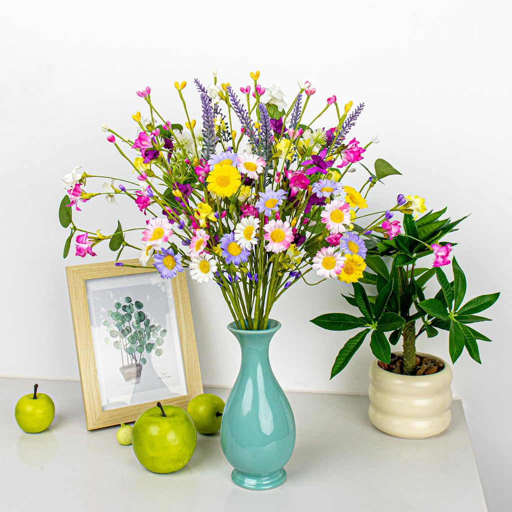 Colorful artificial spring floral arrangement in a teal vase with daisies, wildflowers, and berries, next to a framed picture and green apples.