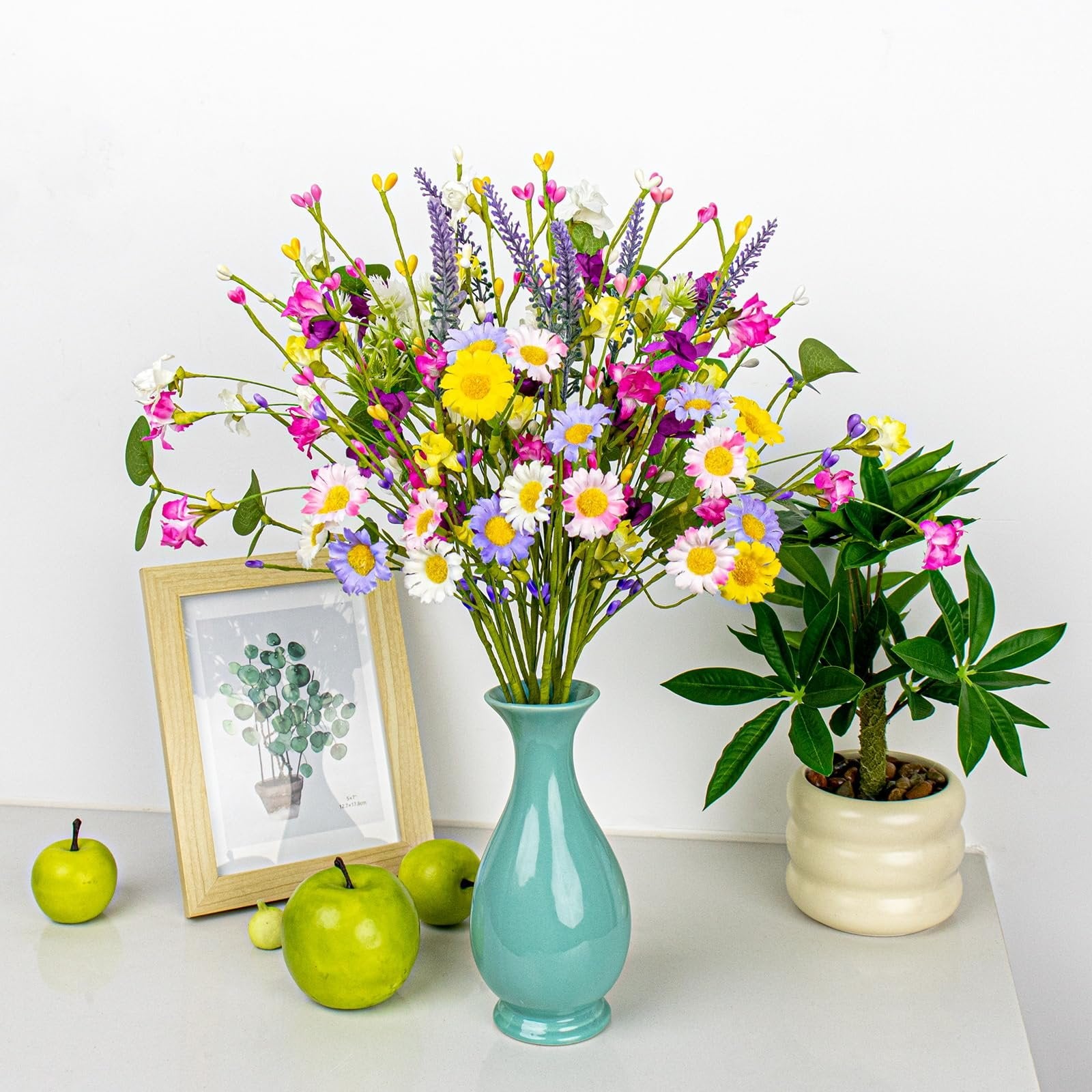 Colorful artificial spring floral arrangement in a teal vase with daisies, wildflowers, and berries, next to a framed picture and green apples.