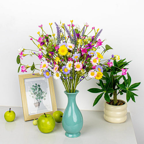 Colorful artificial spring floral arrangement in a teal vase with daisies, wildflowers, and berries, next to a framed picture and green apples.