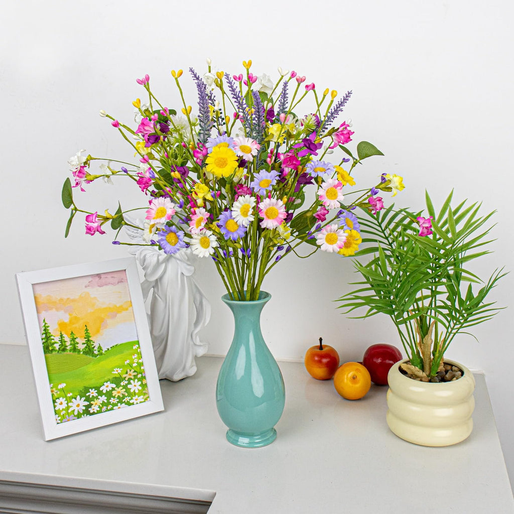 A vibrant bouquet of artificial spring flowers in a teal vase, with a framed landscape painting and a potted plant on a white surface.