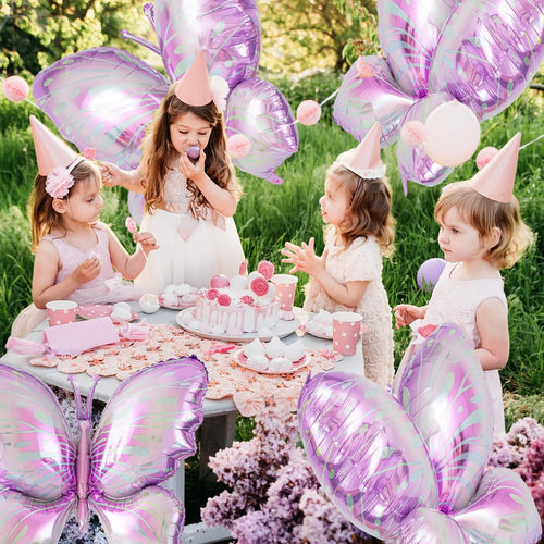 Four young girls in party hats at a table with a cake and balloons, celebrating outdoors.