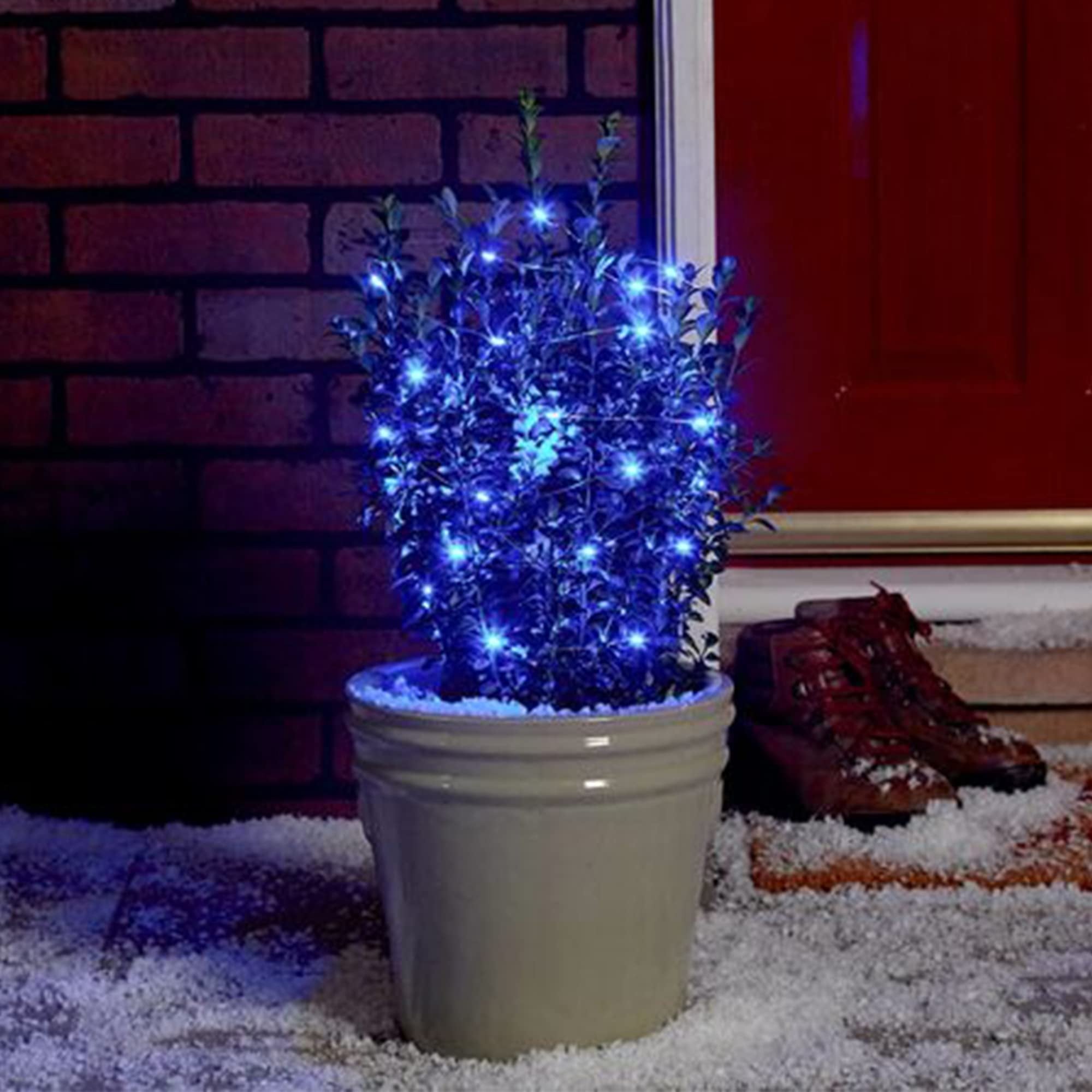 Potted plant decorated with blue LED fairy lights on a snowy porch with red boots nearby