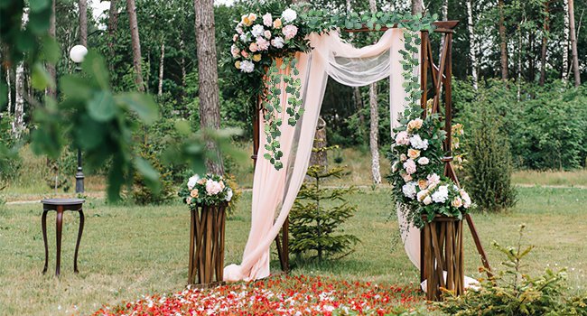 Wedding arch decorated with artificial eucalyptus garland and flowers, set in a grassy outdoor area with trees.