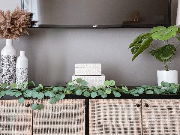 Artificial eucalyptus garland draped across a woven cabinet with decorative vases and a potted plant.