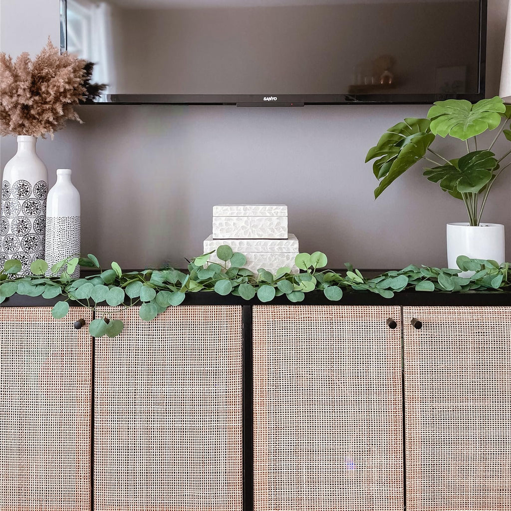 Artificial eucalyptus garland draped across a black cabinet with woven doors, flanked by decorative vases and boxes.