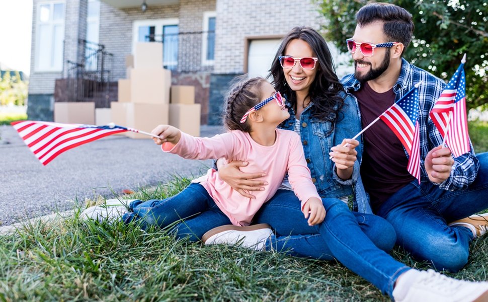 Family with American flags and patriotic sunglasses sitting on grass in front of a house with moving boxes