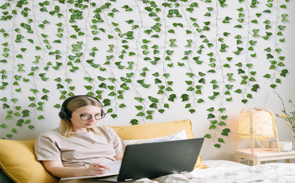 Woman with headphones and glasses working on a laptop, surrounded by artificial ivy vines on a white wall.
