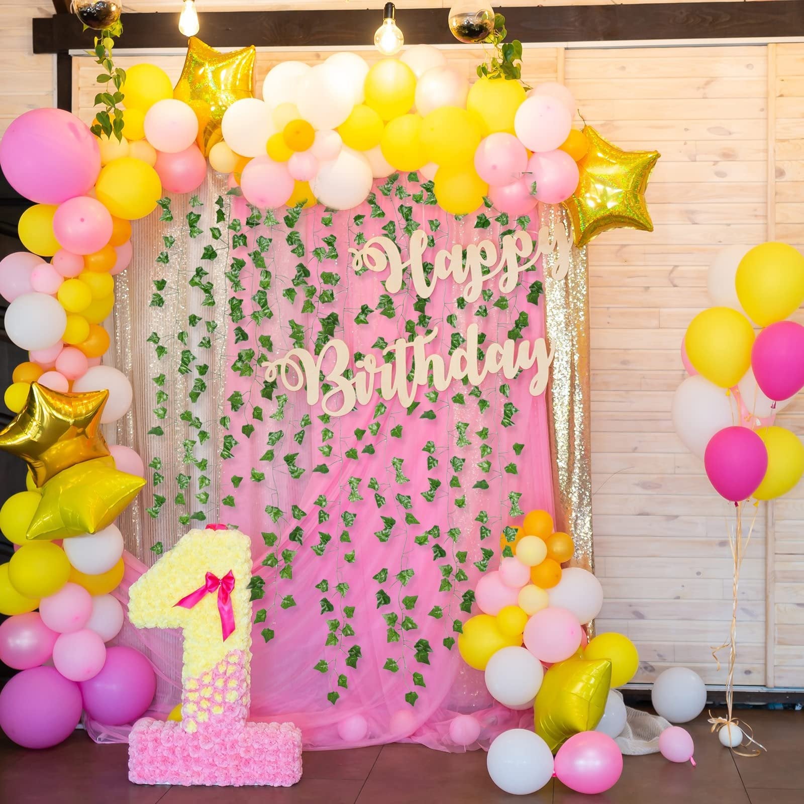 Birthday party backdrop with pink and yellow balloons, artificial ivy vines, and a large number '1' decoration.