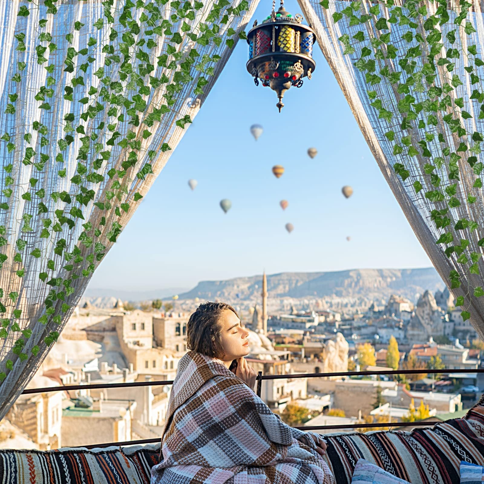 Woman wrapped in blanket on balcony overlooking Cappadocia with hot air balloons, framed by artificial ivy vines.