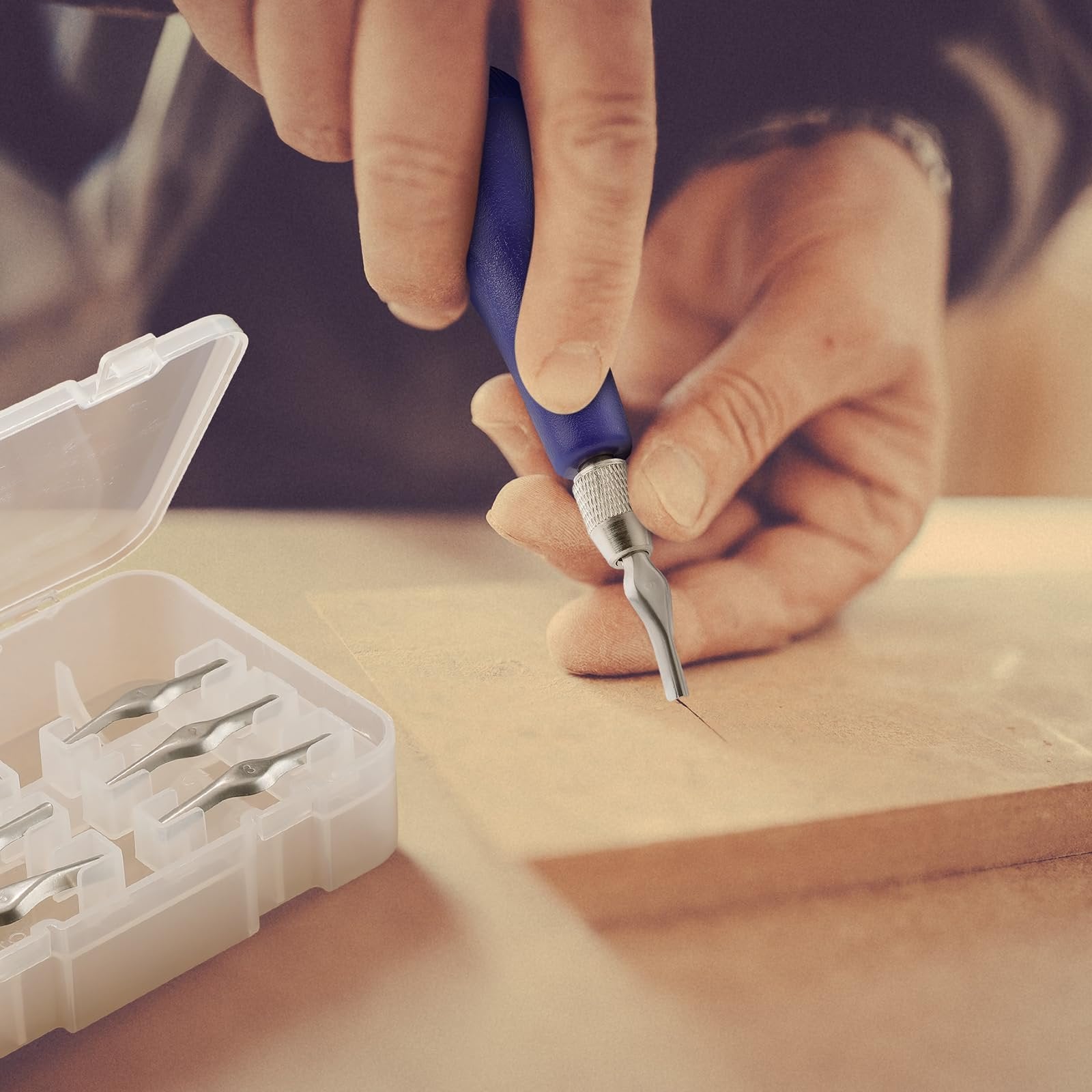Hands using a craft linoleum cutter tool to carve a line into wood, with a case of assorted blades nearby.