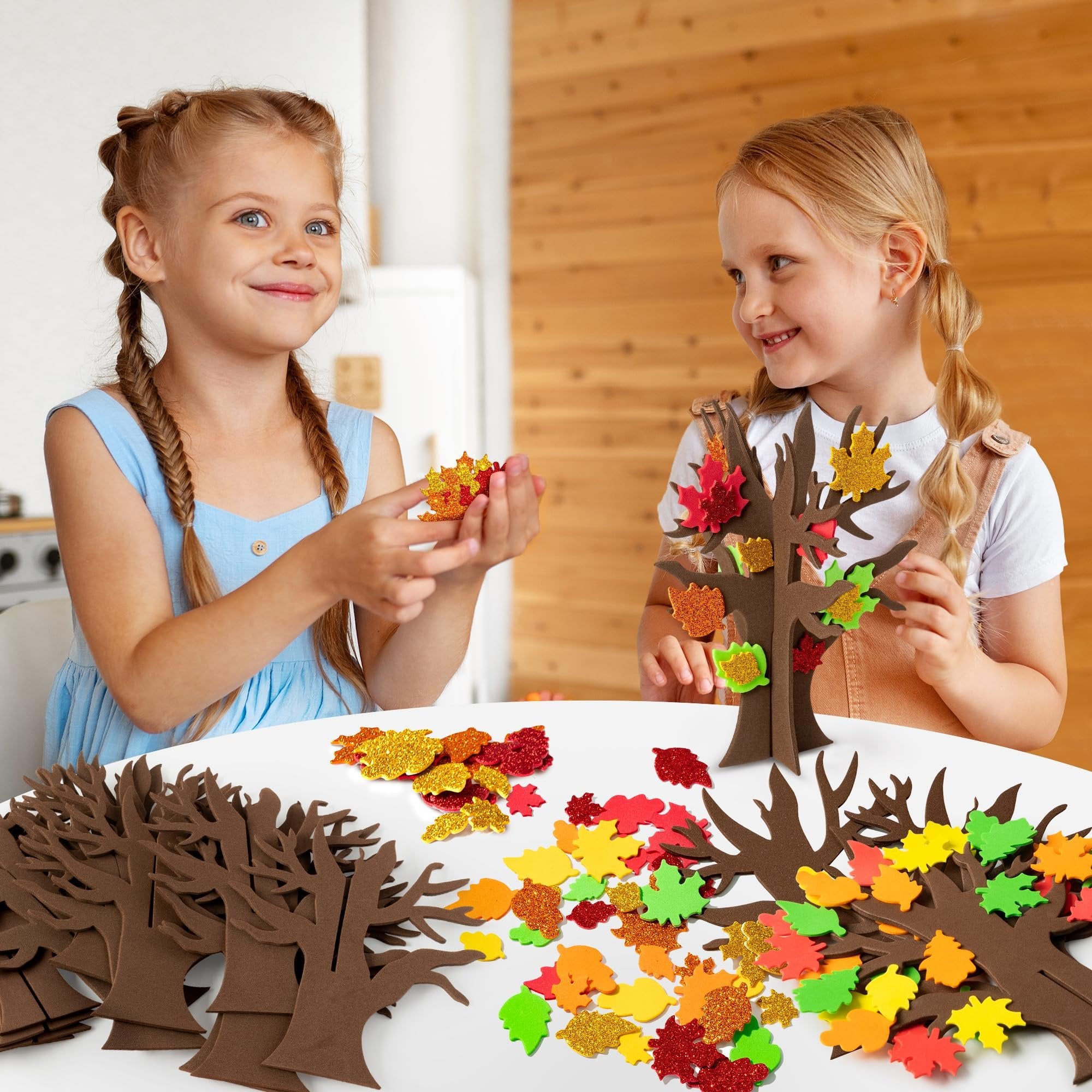 Two young girls crafting fall trees with foam leaves, glitter stickers, and tree bases on a white table.