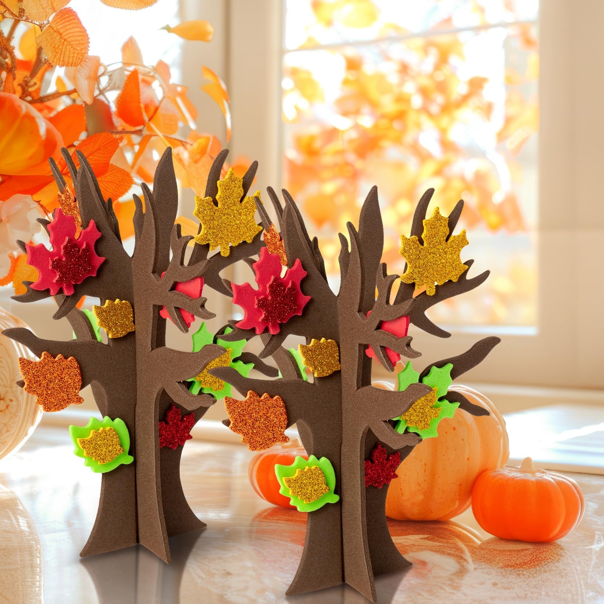 Two foam thankful trees decorated with glitter maple leaf stickers, with pumpkins and fall foliage in the background.