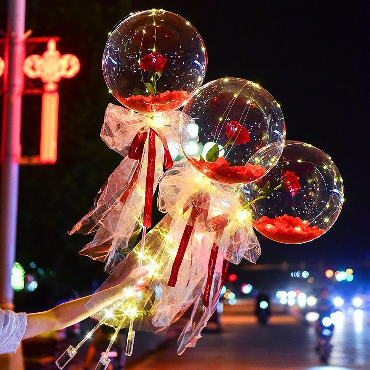 Three clear LED light-up balloons with red roses and warm white string lights held by a hand at night