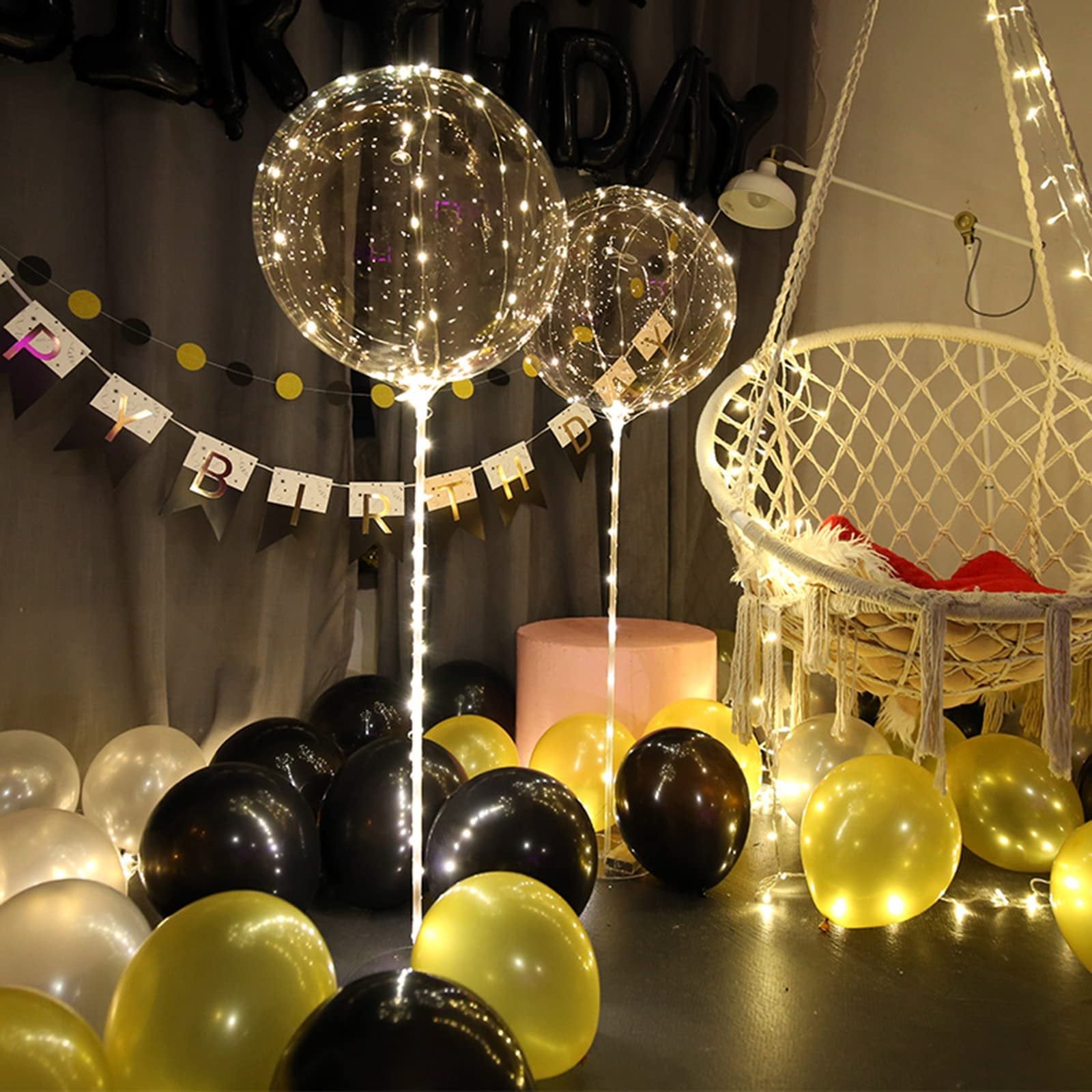 Two clear LED light-up balloons with warm white string lights, surrounded by black, gold, and white balloons, with a macrame chair in the background.