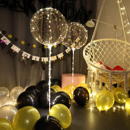 Two clear LED light-up balloons with warm white string lights, surrounded by black, gold, and white balloons, with a macrame chair in the background.