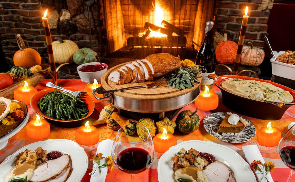 Thanksgiving dinner table with carved turkey, side dishes, pumpkins, and flickering LED tea lights by a fireplace.
