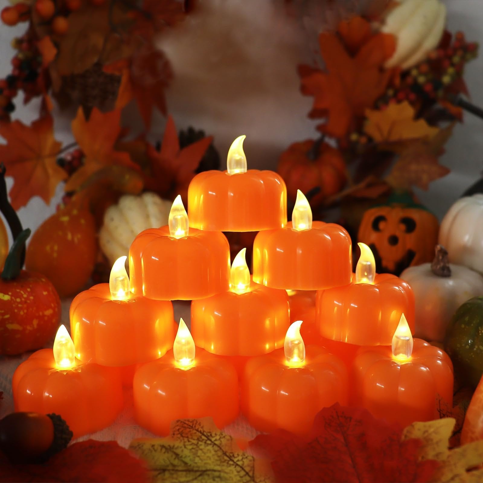 Stack of 12 orange LED pumpkin tea lights with flickering flames, arranged in a pyramid shape with autumn leaves and decorative pumpkins in the background.