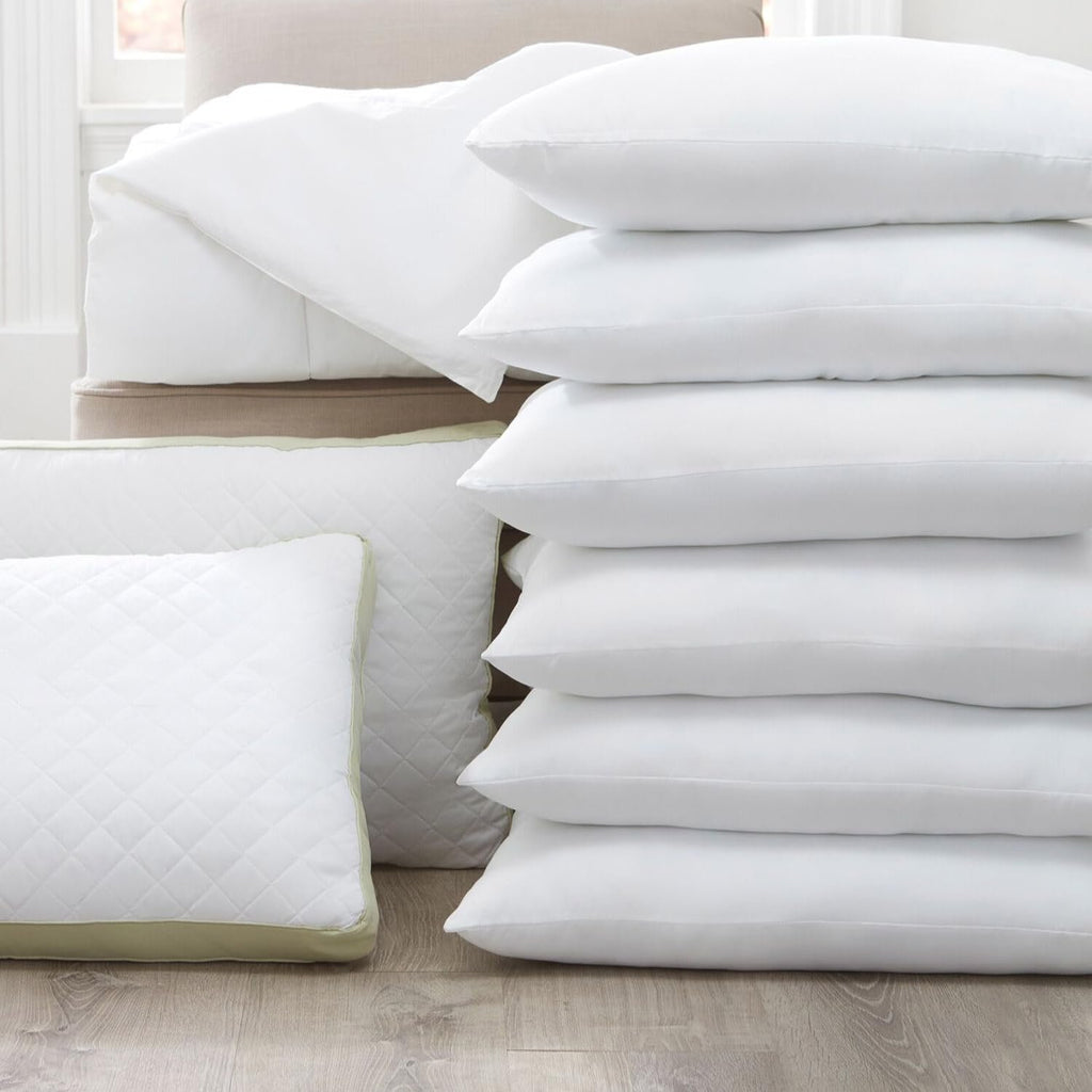 Stack of white microfiber pillowcases with envelope closure and a quilted pillow on a wooden floor.