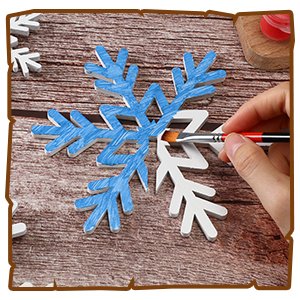 Hand painting a white wooden snowflake decoration with blue strokes on a wooden table, surrounded by other snowflakes.