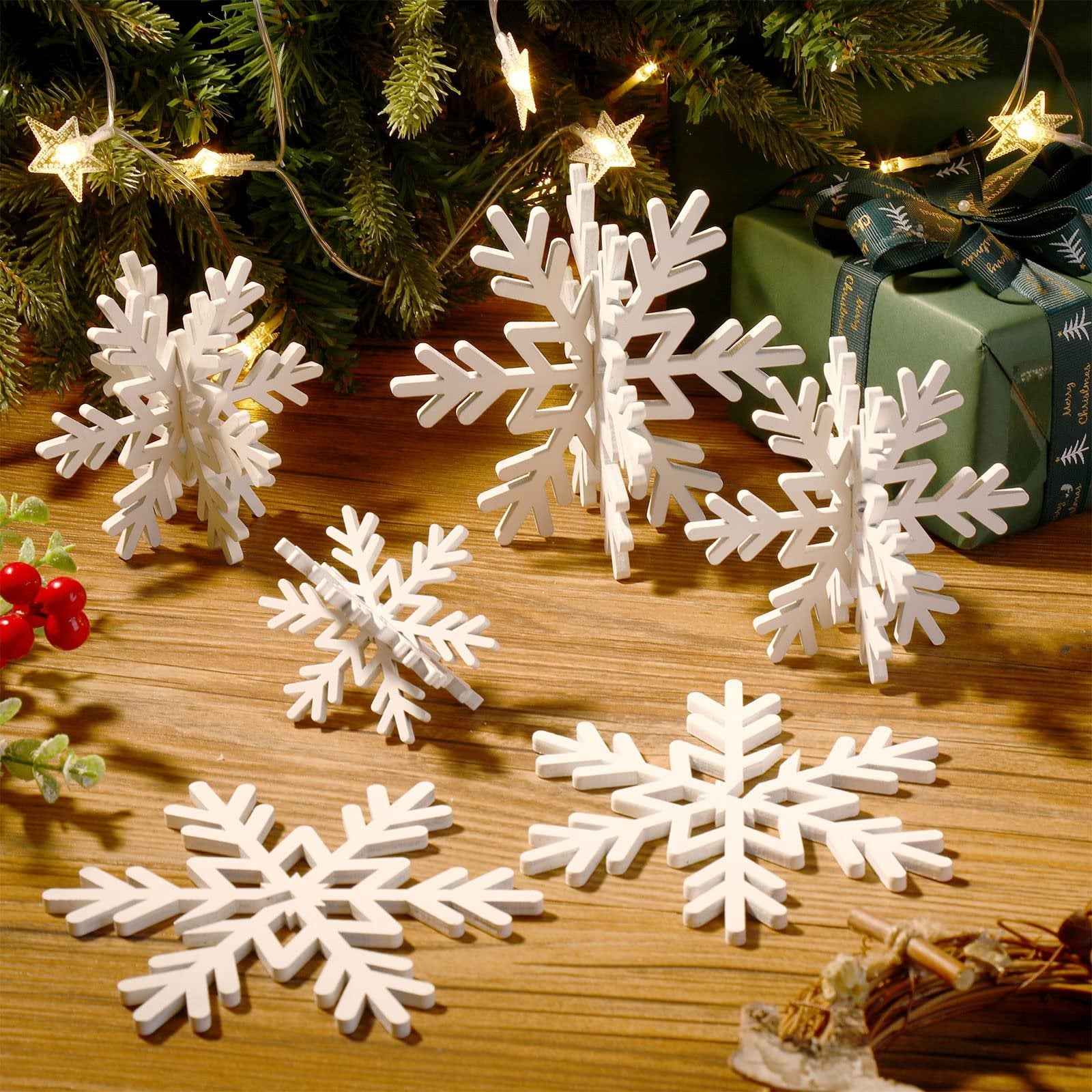 White 3D wooden snowflakes in various sizes arranged on a wooden surface with a Christmas tree and gift in the background.