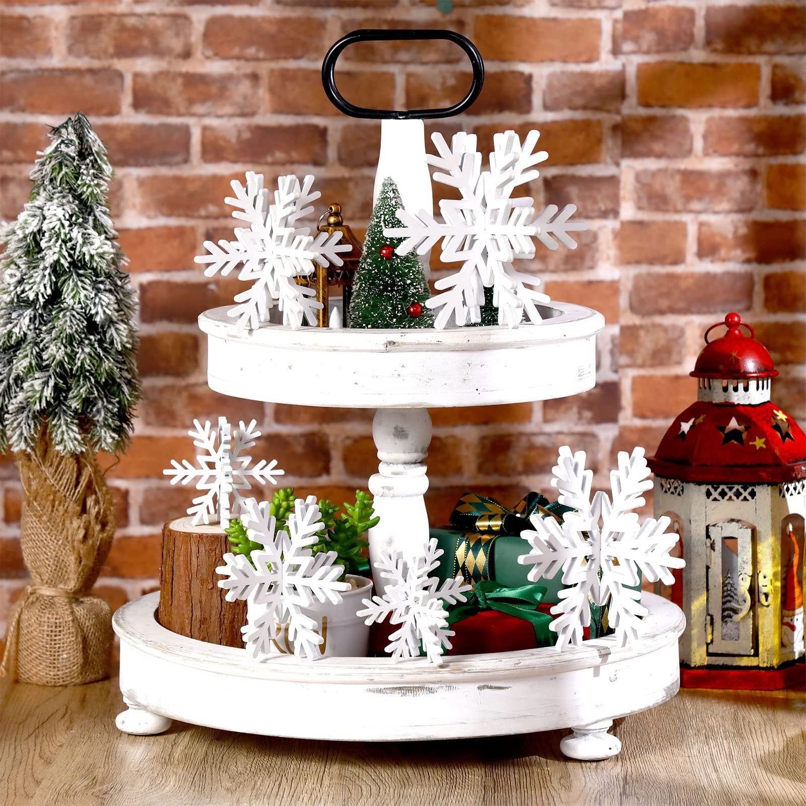 Tiered tray decorated with white wooden snowflakes, mini Christmas trees, gifts, and a lantern against a brick background.